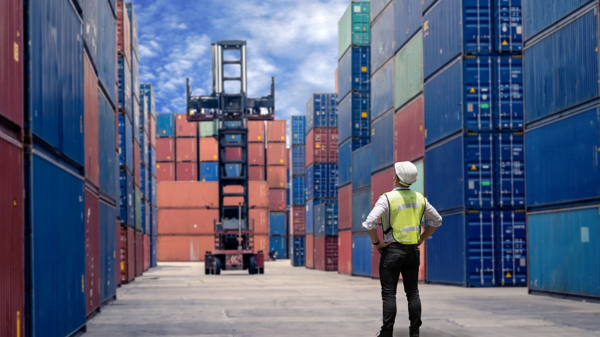 A supervisor in a safety vest watches a reach stacker working between rows of shipping containers.